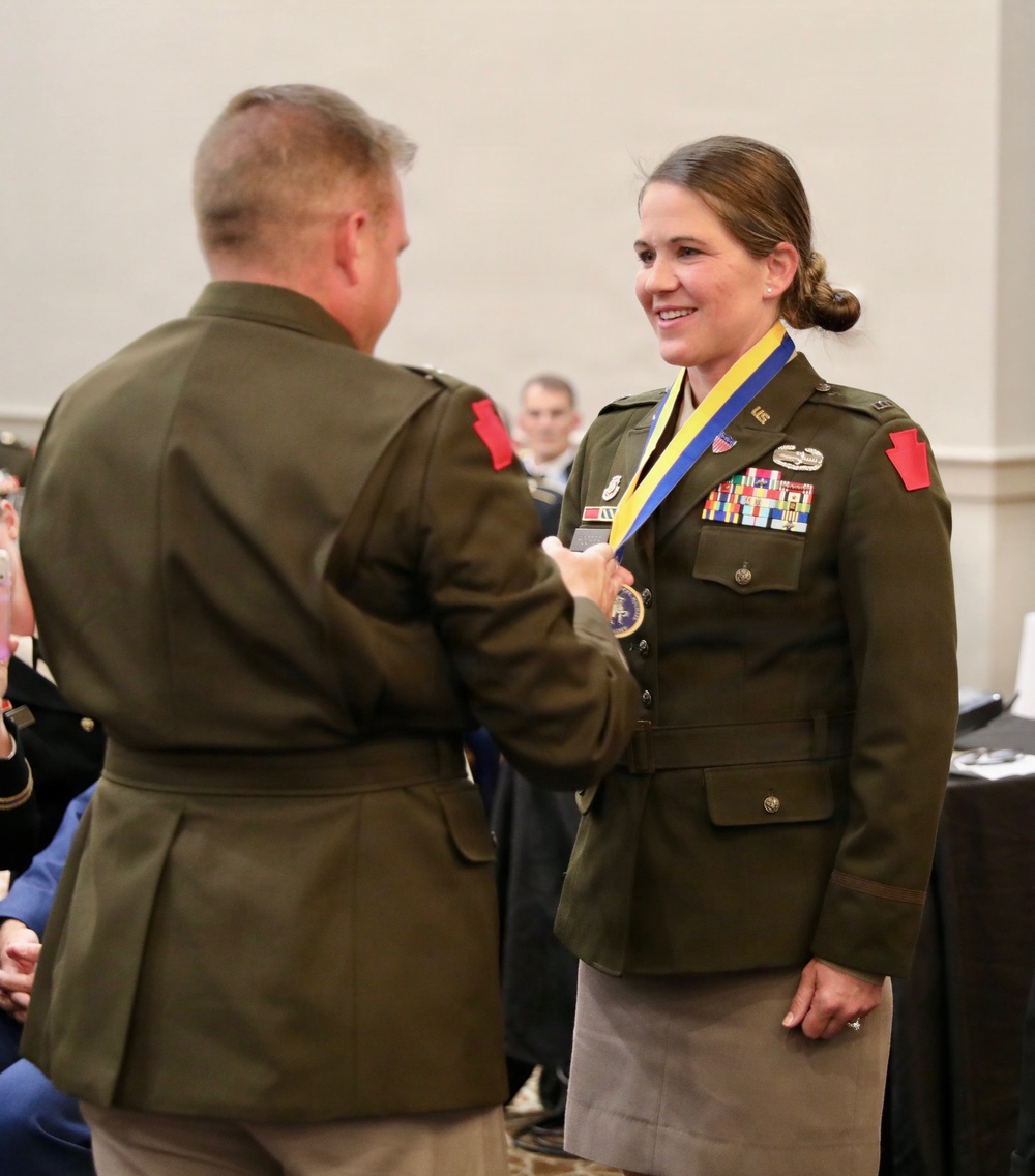 A military ceremony in which a male officer is presenting a medal on a blue and yellow ribbon to a female officer in U.S. Army dress green uniform. The female officer is smiling and wearing multiple service ribbons, badges, and a name tag. The event appears to take place in a formal indoor venue with an audience visible in the background.