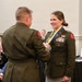 A military ceremony in which a male officer is presenting a medal on a blue and yellow ribbon to a female officer in U.S. Army dress green uniform. The female officer is smiling and wearing multiple service ribbons, badges, and a name tag. The event appears to take place in a formal indoor venue with an audience visible in the background.