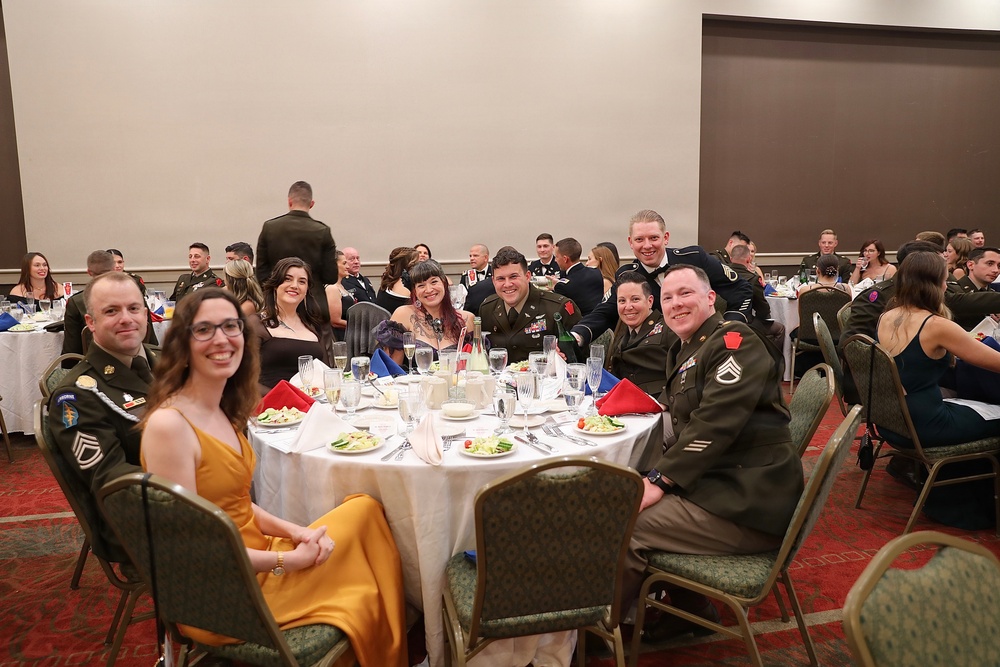 A formal military banquet or ball taking place in a hotel ballroom, with multiple tables of attendees including uniformed U.S. Army soldiers and their civilian guests. The foreground table features a group of approximately eight people posing and smiling for the camera. The event appears to be a formal dining occasion with decorated tables, glassware, and formal attire.