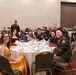 A formal military banquet or ball taking place in a hotel ballroom, with multiple tables of attendees including uniformed U.S. Army soldiers and their civilian guests. The foreground table features a group of approximately eight people posing and smiling for the camera. The event appears to be a formal dining occasion with decorated tables, glassware, and formal attire.