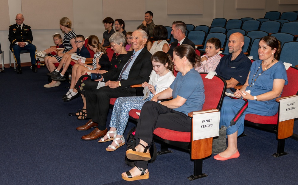 A formal ceremony is taking place in an auditorium or conference room, with family members seated in designated 'Family Seating' sections. The audience includes multiple generations, from young children to elderly individuals, suggesting a military or official recognition event. A uniformed military officer is visible seated to the left side of the room.