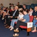 A formal ceremony is taking place in an auditorium or conference room, with family members seated in designated 'Family Seating' sections. The audience includes multiple generations, from young children to elderly individuals, suggesting a military or official recognition event. A uniformed military officer is visible seated to the left side of the room.