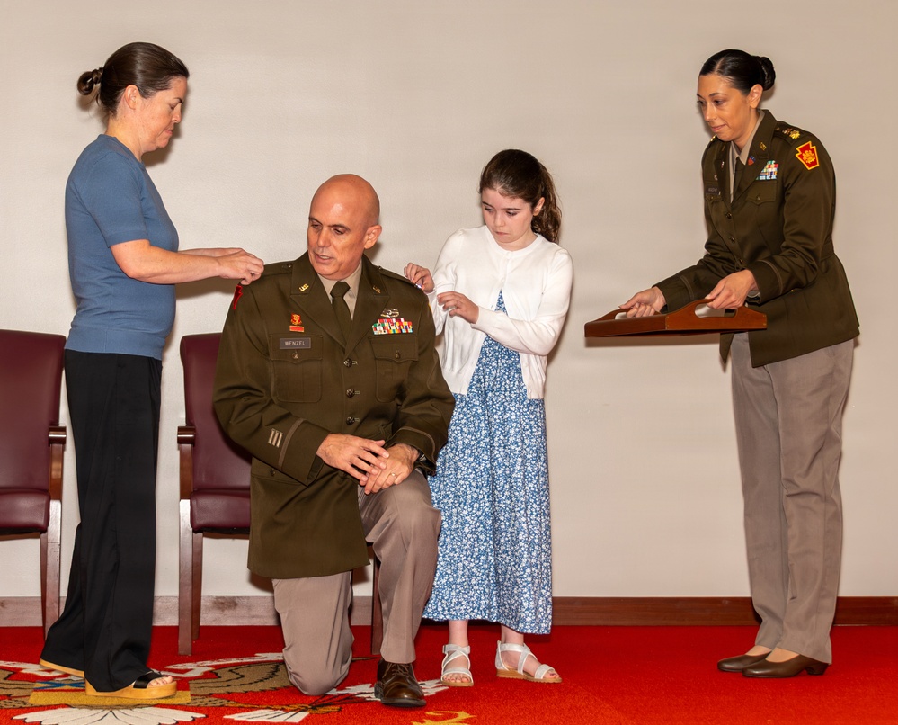A military promotion ceremony is taking place in an indoor setting. A male soldier identified by his name tape as 'WENZEL' is kneeling while a woman and a young girl pin new rank insignia onto his shoulders. A female soldier in uniform assists by holding a presentation tray with the rank devices.