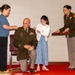 A military promotion ceremony is taking place in an indoor setting. A male soldier identified by his name tape as 'WENZEL' is kneeling while a woman and a young girl pin new rank insignia onto his shoulders. A female soldier in uniform assists by holding a presentation tray with the rank devices.
