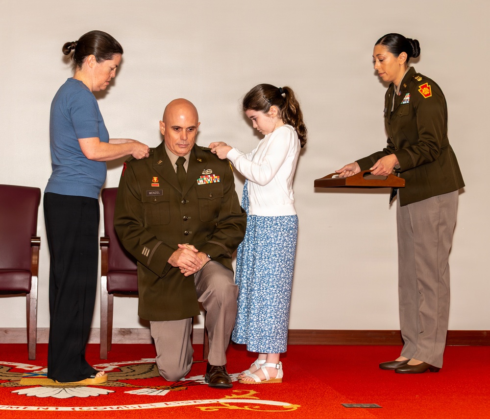 A U.S. Army officer named Wenzel is kneeling during what appears to be a promotion ceremony. A woman (likely his spouse) and a young girl (likely his daughter) are pinning new rank insignia onto his shoulders, while a female Army officer stands nearby at a podium holding items for the ceremony. The event takes place in a formal military setting with a red ceremonial carpet.