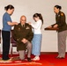 A U.S. Army officer named Wenzel is kneeling during what appears to be a promotion ceremony. A woman (likely his spouse) and a young girl (likely his daughter) are pinning new rank insignia onto his shoulders, while a female Army officer stands nearby at a podium holding items for the ceremony. The event takes place in a formal military setting with a red ceremonial carpet.