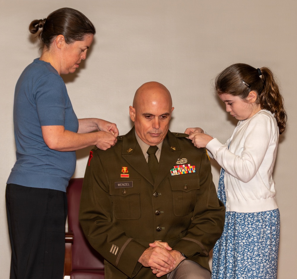 A U.S. Army officer named Wenzel is seated while a woman and a young girl pin new rank insignia onto his shoulders, indicating a military promotion ceremony. The officer wears the Army Service Uniform (green) with multiple service ribbons, a name tag reading 'WENZEL,' and collar insignia. This appears to be a formal promotion pinning ceremony with family members participating.