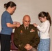 A U.S. Army officer named Wenzel is seated while a woman and a young girl pin new rank insignia onto his shoulders, indicating a military promotion ceremony. The officer wears the Army Service Uniform (green) with multiple service ribbons, a name tag reading 'WENZEL,' and collar insignia. This appears to be a formal promotion pinning ceremony with family members participating.