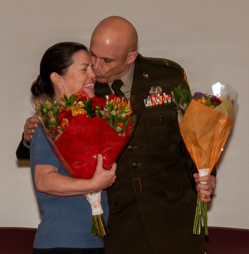 A military officer in dress green uniform is kissing a smiling woman on the cheek during what appears to be a formal ceremony or celebration. The officer holds a bouquet of flowers wrapped in orange tissue paper, while the woman holds a large bouquet of red and mixed flowers. The scene suggests a celebratory military event such as a promotion, retirement, or award ceremony.