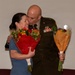A military officer in dress green uniform is kissing a smiling woman on the cheek during what appears to be a formal ceremony or celebration. The officer holds a bouquet of flowers wrapped in orange tissue paper, while the woman holds a large bouquet of red and mixed flowers. The scene suggests a celebratory military event such as a promotion, retirement, or award ceremony.
