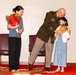 A military officer in dress uniform bends down to embrace and kiss a young girl who is holding a bouquet of flowers. A woman standing to the left also holds a large bouquet of red and orange flowers and smiles warmly. The setting appears to be a formal military ceremony or promotion event, likely involving family members being honored.