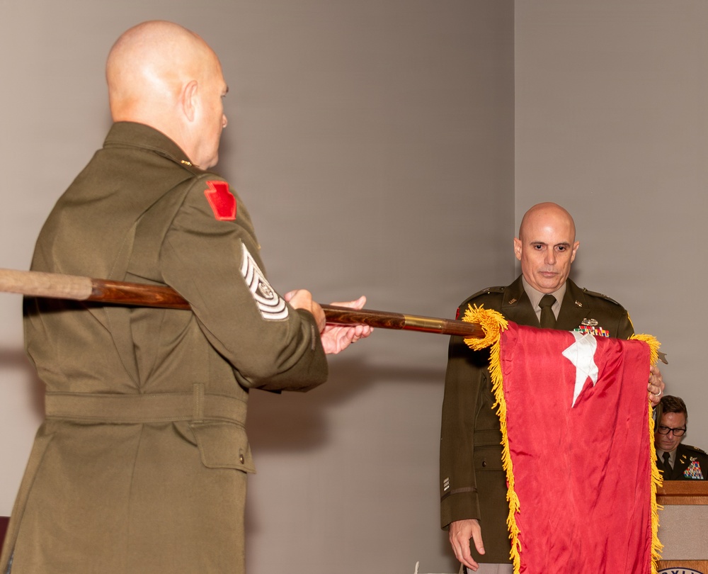 Two U.S. Army soldiers in dress green uniforms are participating in a formal military ceremony, likely a change of command or promotion ceremony. One soldier (facing away) holds a wooden staff while the other holds a red general officer flag with a single white star and gold fringe, indicating a Brigadier General. A third uniformed individual is partially visible in the background near a podium.