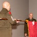 Two U.S. Army soldiers in dress green uniforms are participating in a formal military ceremony, likely a change of command or promotion ceremony. One soldier (facing away) holds a wooden staff while the other holds a red general officer flag with a single white star and gold fringe, indicating a Brigadier General. A third uniformed individual is partially visible in the background near a podium.