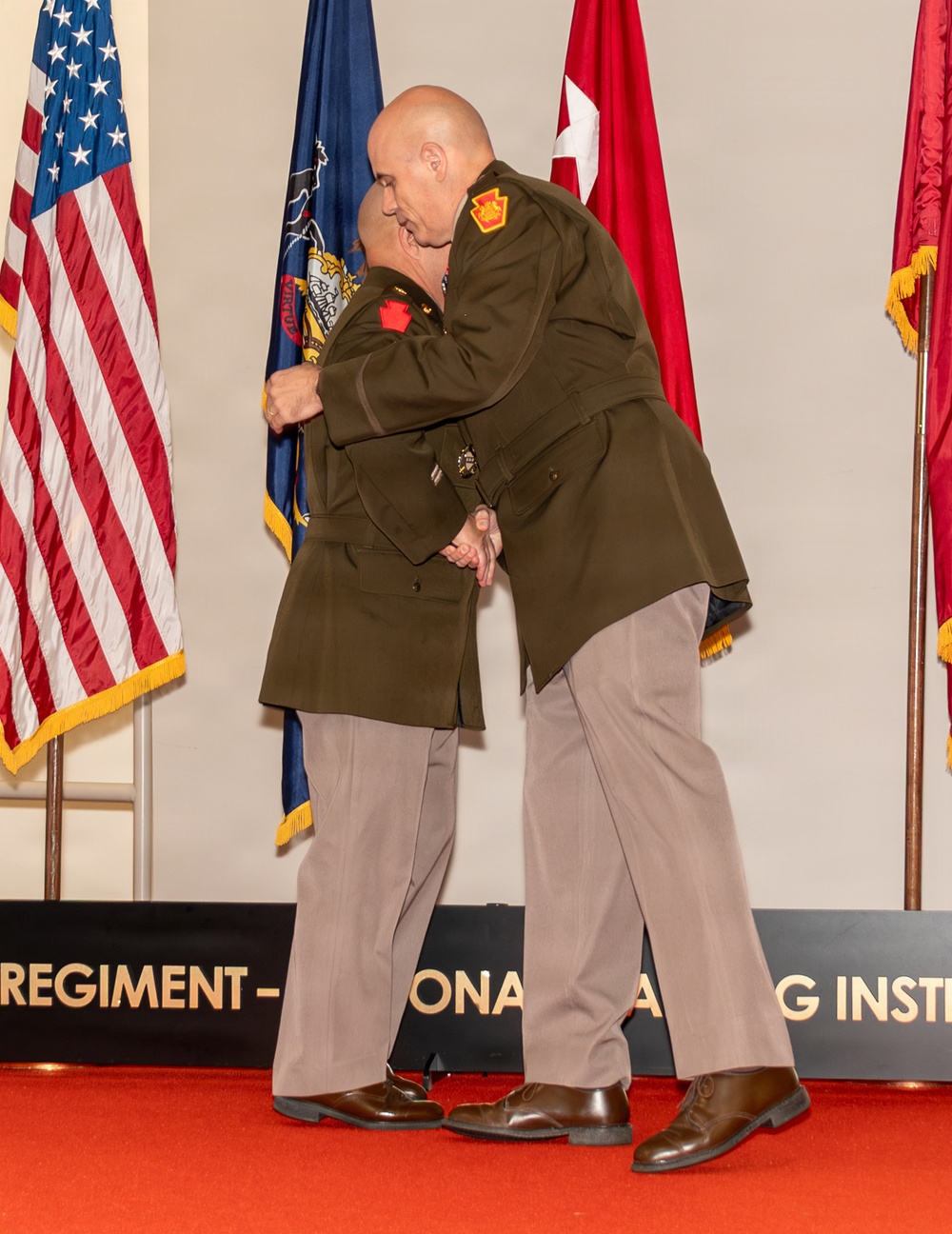 Two uniformed individuals in olive green military-style jackets and tan trousers are embracing on a ceremonial stage with a red carpet. The setting appears to be a formal military or law enforcement ceremony, indicated by the flags and institutional signage visible in the background. Both individuals wear matching uniforms with red and gold shoulder patches.
