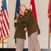 Two uniformed individuals in olive green military-style jackets and tan trousers are embracing on a ceremonial stage with a red carpet. The setting appears to be a formal military or law enforcement ceremony, indicated by the flags and institutional signage visible in the background. Both individuals wear matching uniforms with red and gold shoulder patches.