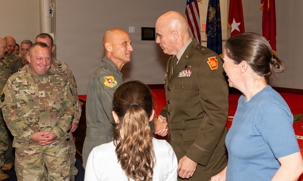 A military ceremony or reception is taking place in a formal indoor setting with flags displayed in the background. Two senior military officers are shaking hands at the center of the image, while other personnel and civilians look on. The setting appears to be an official military function, possibly a promotion, retirement, or change of command ceremony.