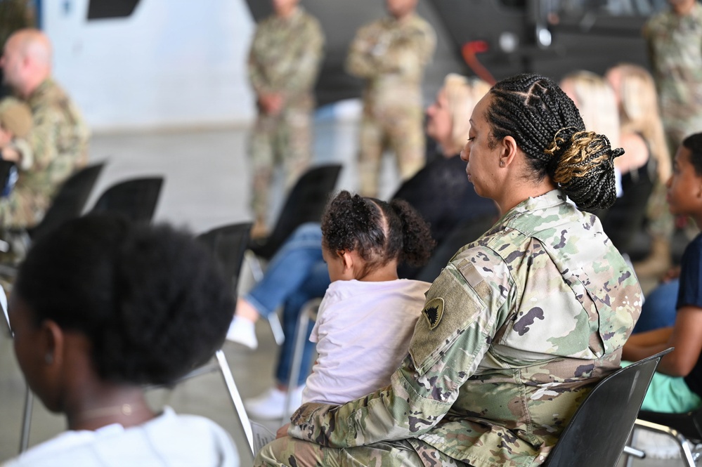 A military gathering or briefing is taking place in what appears to be a hangar or large indoor facility. A female soldier in OCP (Operational Camouflage Pattern) uniform is seated in the foreground, holding a young child on her lap. The audience consists of a mix of uniformed military personnel and civilians, with additional soldiers standing in the background.