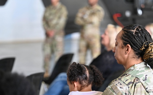 A military gathering or briefing is taking place in what appears to be a hangar or large indoor facility. A female soldier in OCP (Operational Camouflage Pattern) uniform is seated in the foreground, holding a young child on her lap. The audience consists of a mix of uniformed military personnel and civilians, with additional soldiers standing in the background.