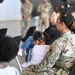 A military gathering or briefing is taking place in what appears to be a hangar or large indoor facility. A female soldier in OCP (Operational Camouflage Pattern) uniform is seated in the foreground, holding a young child on her lap. The audience consists of a mix of uniformed military personnel and civilians, with additional soldiers standing in the background.