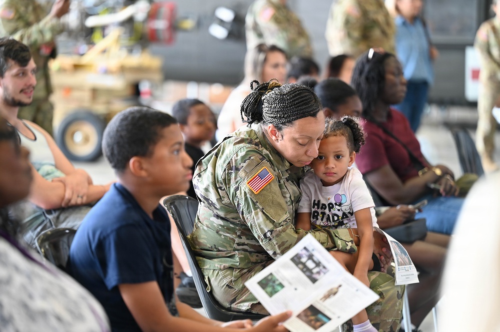 A U.S. Army soldier in MultiCam/OCP camouflage uniform is seated in a hangar setting, holding and nuzzling a young child wearing a 'Happy Birthday' shirt. The scene appears to be a military family event or unit ceremony, with multiple civilians, family members, and other service members visible in the background. The soldier is also holding a printed program or informational booklet.