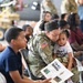 A U.S. Army soldier in MultiCam/OCP camouflage uniform is seated in a hangar setting, holding and nuzzling a young child wearing a 'Happy Birthday' shirt. The scene appears to be a military family event or unit ceremony, with multiple civilians, family members, and other service members visible in the background. The soldier is also holding a printed program or informational booklet.