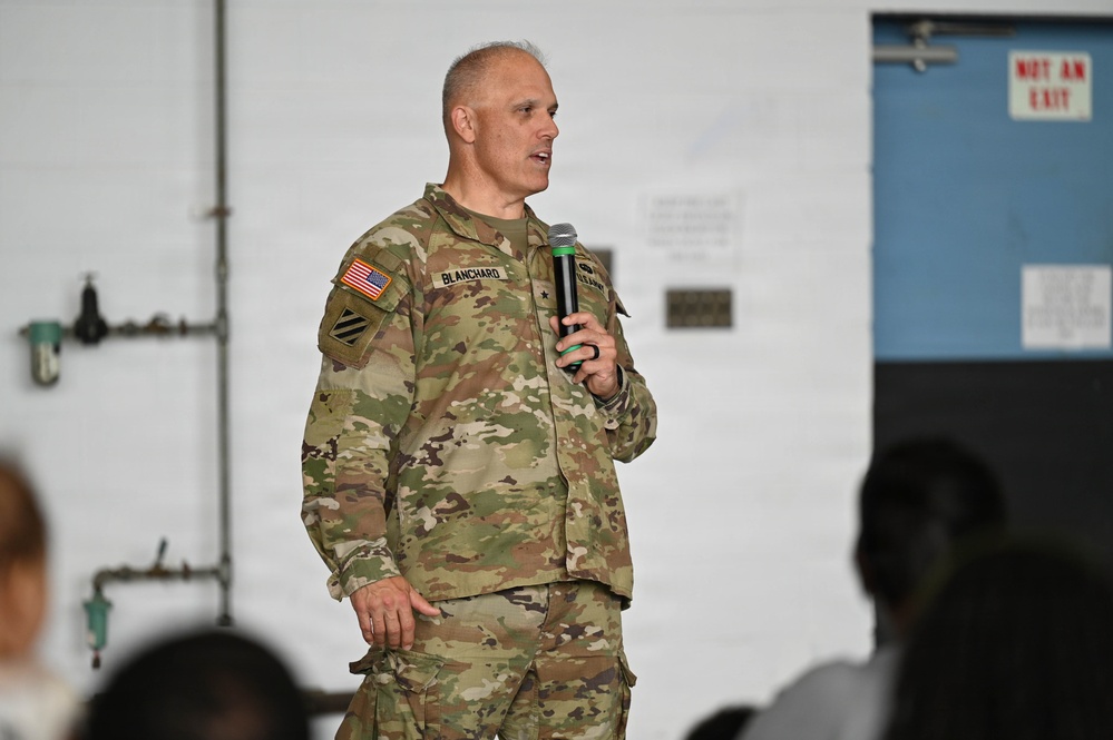 A U.S. Army officer identified by name tape as 'BLANCHARD' is speaking into a microphone at what appears to be a military briefing or town hall event. He is wearing the Operational Camouflage Pattern (OCP) uniform with a U.S. flag patch, 3rd Infantry Division patch, and a general officer star visible on his chest. An audience is partially visible in the foreground as silhouettes.