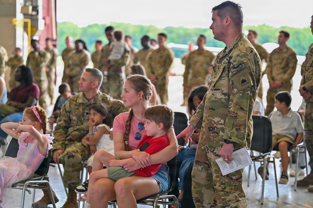 A military ceremony or formation event taking place inside what appears to be an aircraft hangar, with numerous soldiers in OCP (Operational Camouflage Pattern) uniforms present alongside civilian family members. A woman in a pink shirt holds a young child while a standing soldier in uniform appears to interact with them. The scene suggests a unit event such as a deployment ceremony, homecoming, or change of command.