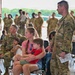 A military ceremony or formation event taking place inside what appears to be an aircraft hangar, with numerous soldiers in OCP (Operational Camouflage Pattern) uniforms present alongside civilian family members. A woman in a pink shirt holds a young child while a standing soldier in uniform appears to interact with them. The scene suggests a unit event such as a deployment ceremony, homecoming, or change of command.