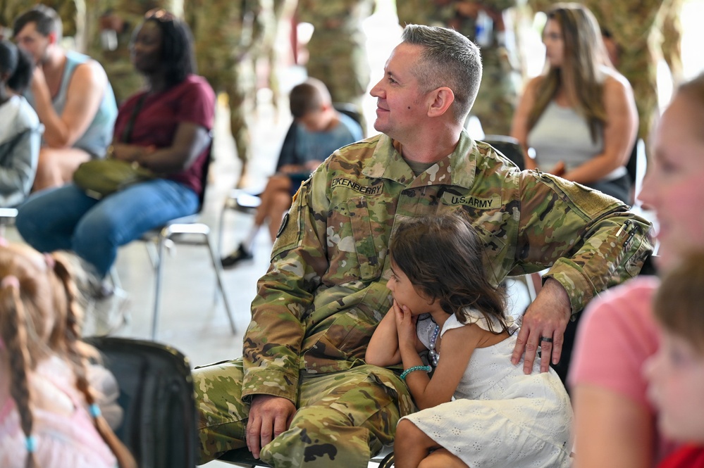 A U.S. Army soldier named Eikenberry, wearing Operational Camouflage Pattern (OCP) uniform, sits with a young girl on his lap during what appears to be a military family event or ceremony. The soldier is smiling and looking to the side while the child rests against him. The setting appears to be an outdoor or semi-covered gathering with multiple families and other military personnel present.