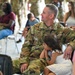 A U.S. Army soldier named Eikenberry, wearing Operational Camouflage Pattern (OCP) uniform, sits with a young girl on his lap during what appears to be a military family event or ceremony. The soldier is smiling and looking to the side while the child rests against him. The setting appears to be an outdoor or semi-covered gathering with multiple families and other military personnel present.