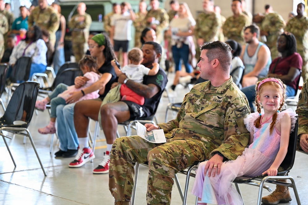 A U.S. Army soldier identified by the name tape 'SPRINGER' sits with a young red-haired girl in a pink dress during what appears to be a military ceremony or formation event held indoors, likely a hangar or large assembly hall. Multiple soldiers in OCP (Operational Camouflage Pattern) uniforms are present alongside civilians and family members seated in folding chairs. The setting suggests a unit event such as a deployment ceremony, homecoming, or family day.