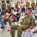 A U.S. Army soldier identified by the name tape 'SPRINGER' sits with a young red-haired girl in a pink dress during what appears to be a military ceremony or formation event held indoors, likely a hangar or large assembly hall. Multiple soldiers in OCP (Operational Camouflage Pattern) uniforms are present alongside civilians and family members seated in folding chairs. The setting suggests a unit event such as a deployment ceremony, homecoming, or family day.