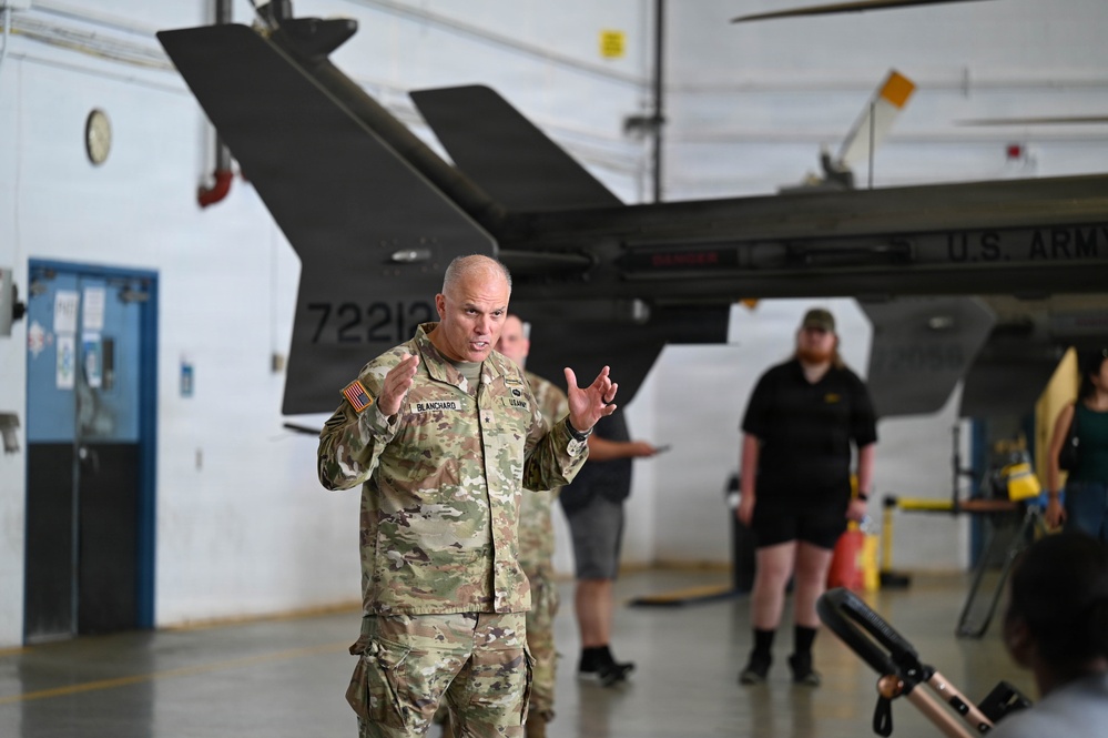 A U.S. Army soldier identified by name tape as 'BLANCHARD' is speaking or briefing in what appears to be a military aircraft hangar. A U.S. Army helicopter (tail number partially visible as 72212) is visible in the background. Several civilians and military personnel are present in the hangar setting.