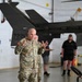 A U.S. Army soldier identified by name tape as 'BLANCHARD' is speaking or briefing in what appears to be a military aircraft hangar. A U.S. Army helicopter (tail number partially visible as 72212) is visible in the background. Several civilians and military personnel are present in the hangar setting.