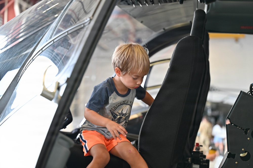 A young blonde boy, approximately 4-6 years old, is climbing into or exploring the cockpit of a helicopter, likely at an aviation museum or air show. He is wearing a gray graphic t-shirt and orange shorts, and appears engaged and curious as he navigates the seat area. The setting appears to be a large hangar with other visitors visible in the background.