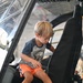 A young blonde boy, approximately 4-6 years old, is climbing into or exploring the cockpit of a helicopter, likely at an aviation museum or air show. He is wearing a gray graphic t-shirt and orange shorts, and appears engaged and curious as he navigates the seat area. The setting appears to be a large hangar with other visitors visible in the background.