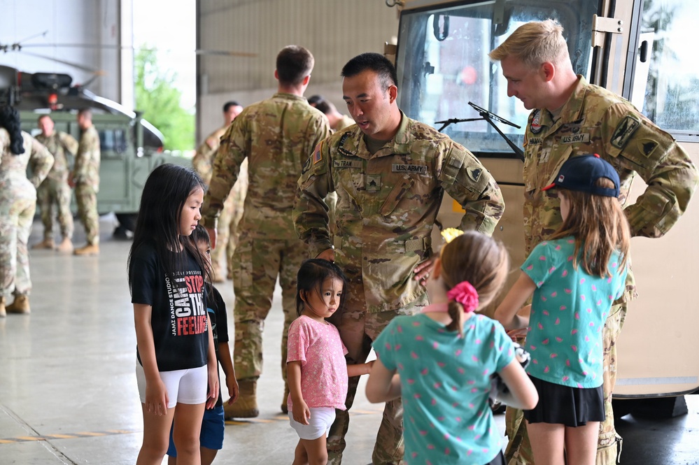 U.S. Army soldiers in OCP (Operational Camouflage Pattern) uniforms interact with young children inside what appears to be a military hangar facility. The central soldier, whose name tape reads 'TRAN,' is bending down to engage with several children, while another soldier stands nearby. The setting suggests a family day, homecoming, or community outreach event at a military installation.