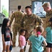 U.S. Army soldiers in OCP (Operational Camouflage Pattern) uniforms interact with young children inside what appears to be a military hangar facility. The central soldier, whose name tape reads 'TRAN,' is bending down to engage with several children, while another soldier stands nearby. The setting suggests a family day, homecoming, or community outreach event at a military installation.