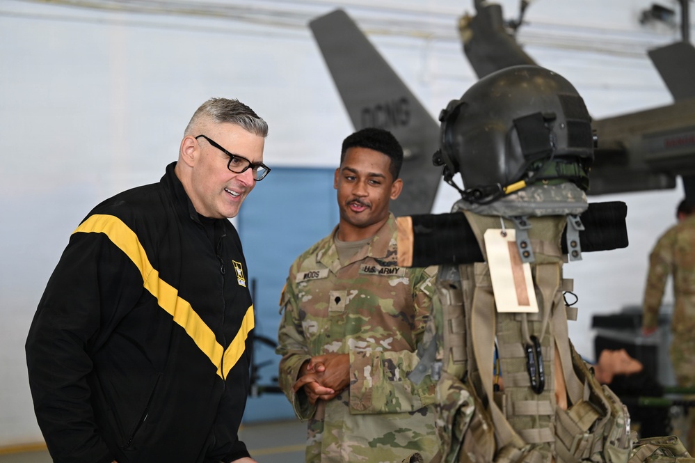 Two individuals are engaged in conversation inside what appears to be a military hangar. One person is wearing a black U.S. Army physical fitness jacket, while the other is in Army Combat Uniform (ACU) with the name tape 'WOODS' visible. A military flight suit or gear display with a helmet is positioned in the foreground.