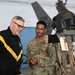 Two individuals are engaged in conversation inside what appears to be a military hangar. One person is wearing a black U.S. Army physical fitness jacket, while the other is in Army Combat Uniform (ACU) with the name tape 'WOODS' visible. A military flight suit or gear display with a helmet is positioned in the foreground.