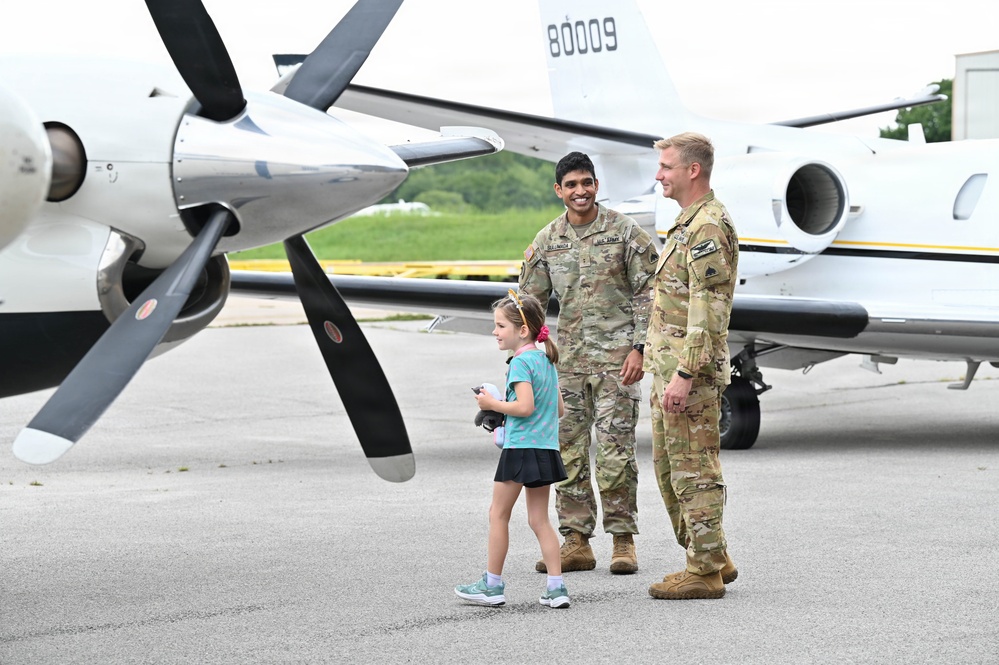 Two military personnel in OCP (Operational Camouflage Pattern) uniforms stand on an airport tarmac alongside a young girl. They appear to be at an airshow or military aviation event, with propeller and jet aircraft visible in the background. The atmosphere is relaxed and friendly, with the soldiers smiling.