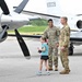 Two military personnel in OCP (Operational Camouflage Pattern) uniforms stand on an airport tarmac alongside a young girl. They appear to be at an airshow or military aviation event, with propeller and jet aircraft visible in the background. The atmosphere is relaxed and friendly, with the soldiers smiling.