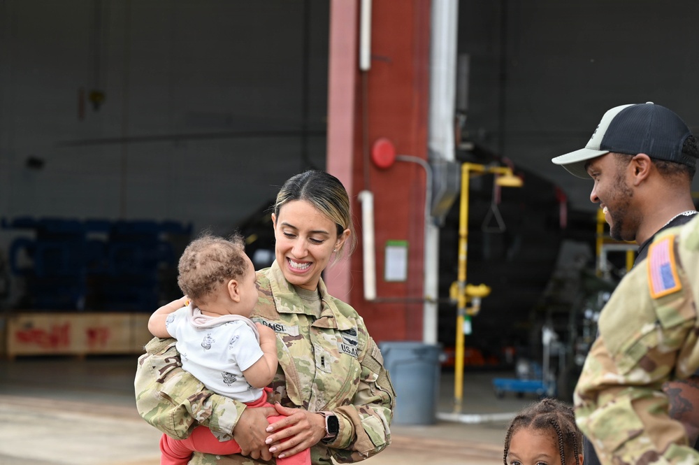A female U.S. Army soldier in OCP (Operational Camouflage Pattern) uniform is smiling warmly while holding an infant, appearing to be in a reunion or family gathering setting near a military hangar. A male soldier in the same uniform stands to the right, also smiling, with a young child partially visible at the lower right. The setting appears to be a military aviation facility based on the hangar and aircraft visible in the background.