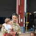 A female U.S. Army soldier in OCP (Operational Camouflage Pattern) uniform is smiling warmly while holding an infant, appearing to be in a reunion or family gathering setting near a military hangar. A male soldier in the same uniform stands to the right, also smiling, with a young child partially visible at the lower right. The setting appears to be a military aviation facility based on the hangar and aircraft visible in the background.
