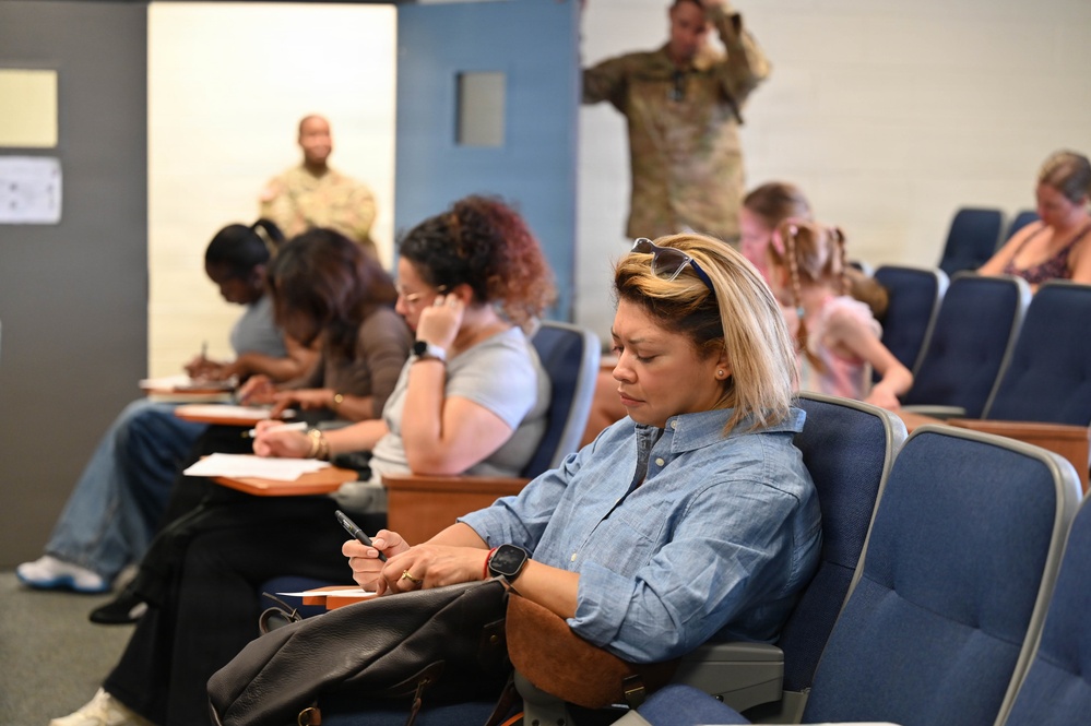 A group of civilians are seated in auditorium-style chairs in what appears to be a briefing or waiting room setting at a military facility. Two individuals in military camouflage uniforms are visible in the background. The civilians appear to be filling out paperwork or using mobile devices while waiting.