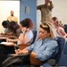A group of civilians are seated in auditorium-style chairs in what appears to be a briefing or waiting room setting at a military facility. Two individuals in military camouflage uniforms are visible in the background. The civilians appear to be filling out paperwork or using mobile devices while waiting.