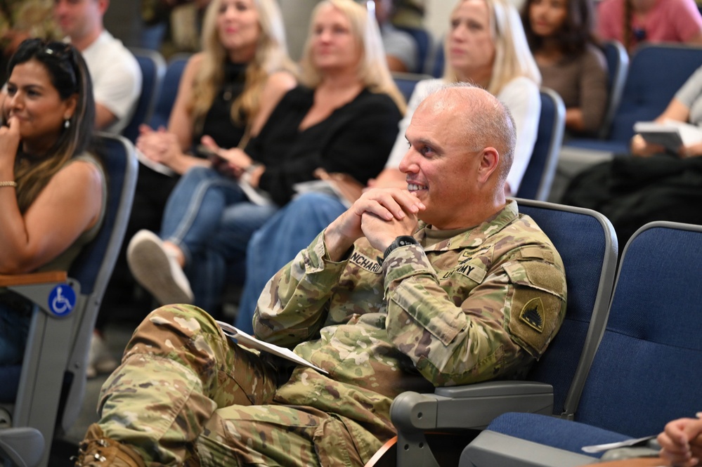 A U.S. Army soldier in OCP (Operational Camouflage Pattern) uniform is seated in an auditorium-style venue, smiling and appearing engaged in a presentation or event. The soldier's name tape reads 'NCHARD' (partially visible, likely 'BLANCHARD' or similar). The audience behind includes several civilians seated in blue auditorium chairs.