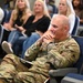 A U.S. Army soldier in OCP (Operational Camouflage Pattern) uniform is seated in an auditorium-style venue, smiling and appearing engaged in a presentation or event. The soldier's name tape reads 'NCHARD' (partially visible, likely 'BLANCHARD' or similar). The audience behind includes several civilians seated in blue auditorium chairs.