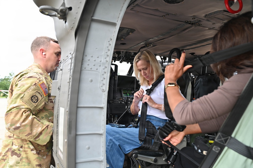 A U.S. Army soldier assigned to the 82nd Airborne Division stands outside a military helicopter, likely a UH-60 Black Hawk, while a civilian woman in a white shirt and jeans fastens her seatbelt in the passenger seat. A second civilian, partially visible on the right, appears to be assisting or observing. The scene suggests a familiarization or orientation visit to a military aircraft.