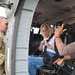 A U.S. Army soldier assigned to the 82nd Airborne Division stands outside a military helicopter, likely a UH-60 Black Hawk, while a civilian woman in a white shirt and jeans fastens her seatbelt in the passenger seat. A second civilian, partially visible on the right, appears to be assisting or observing. The scene suggests a familiarization or orientation visit to a military aircraft.